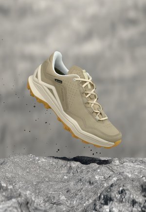 Chaussure de randonnée beige avec une semelle robuste, suspendue au-dessus d'un terrain rocheux, montrant des particules de terre soulevées derrière.