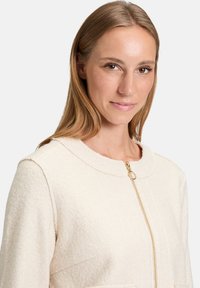 Kremowa polarowa kurtka z okrągłym kołnierzem i złotym zamkiem, z dwiema przednimi kieszeniami i miękką teksturą.