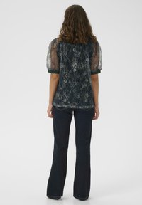 Frau mit lockigem braunem Haar, die ein dunkles Blumenspitzenoberteil mit transparenten Ärmeln und dunklen Schlagjeans trägt, steht vor einem schlichten weißen Hintergrund.