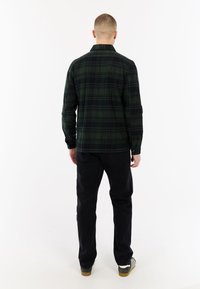 Man die met de rug naar de camera staat, gekleed in een donkergroene en zwarte geblokte shirt, zwarte broek en gestreepte sneakers tegen een witte achtergrond.