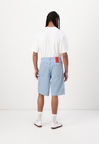 Light blue denim shorts with a textured pattern, five pockets, red branded tag on waistband, paired with a plain white t-shirt and white sneakers.