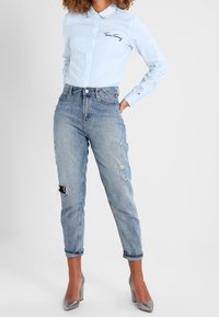 Light blue button-up shirt with an embroidered logo, paired with faded blue jeans featuring distressed areas and rolled cuffs, complemented by silver high heels.