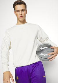 Krämfärgad långärmad Nike-tröja med en enkel logotyp, tillsammans med lila basketshorts med en gul accent, hållande en svart och grå basketboll.