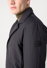 Chaqueta oscura y texturizada con cuello de muesca y un parche de logo redondo en la manga izquierda. Combinada con un suéter de cuello alto en verde oliva por debajo.