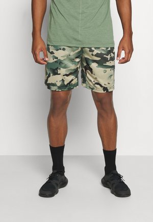 Homme portant un t-shirt vert, un short camouflage vert et beige, des chaussettes noires et des chaussures de sport noires, debout sur un sol gris.