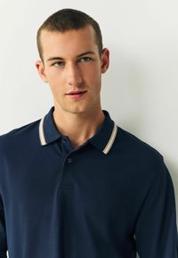 Marineblaues Poloshirt aus strukturiertem Material, mit einem kontrastierenden gestreiften Kragen in Beige und Creme und einem kleinen Logo auf der Brust.