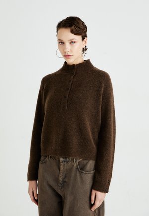 Kvinne iført store hoopsølvøredobber, en brun, fluffy knappegenser og løse, høy midje-brune bukser som står mot en enkel bakgrunn.