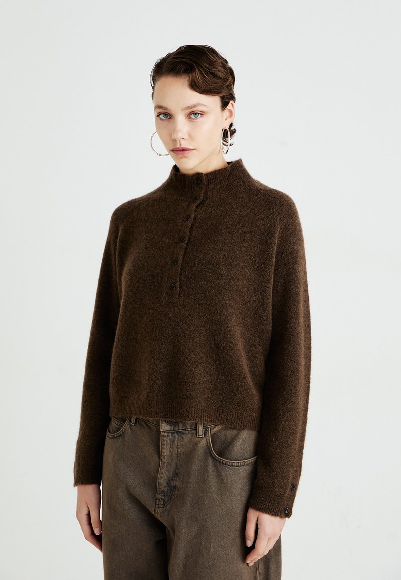 Woman wearing large hoop earrings, brown fuzzy button-up sweater, and loose high-waisted brown pants standing against plain background.