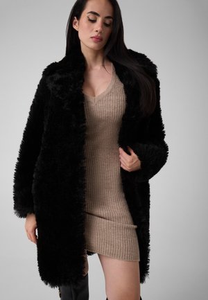 Une femme porte un manteau de fourrure noir sur une robe en maille côtelée couleur taupe, devant un fond gris uni de studio.