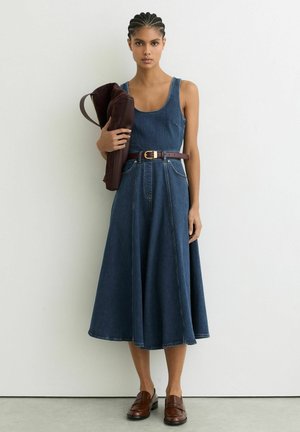 Žena s pletenými vlasmi má na sebe bezrukávové rifľové midi šaty a hnedý opasok, drží zloženú hnedú koženú tašku, stojí pri bielej stene.