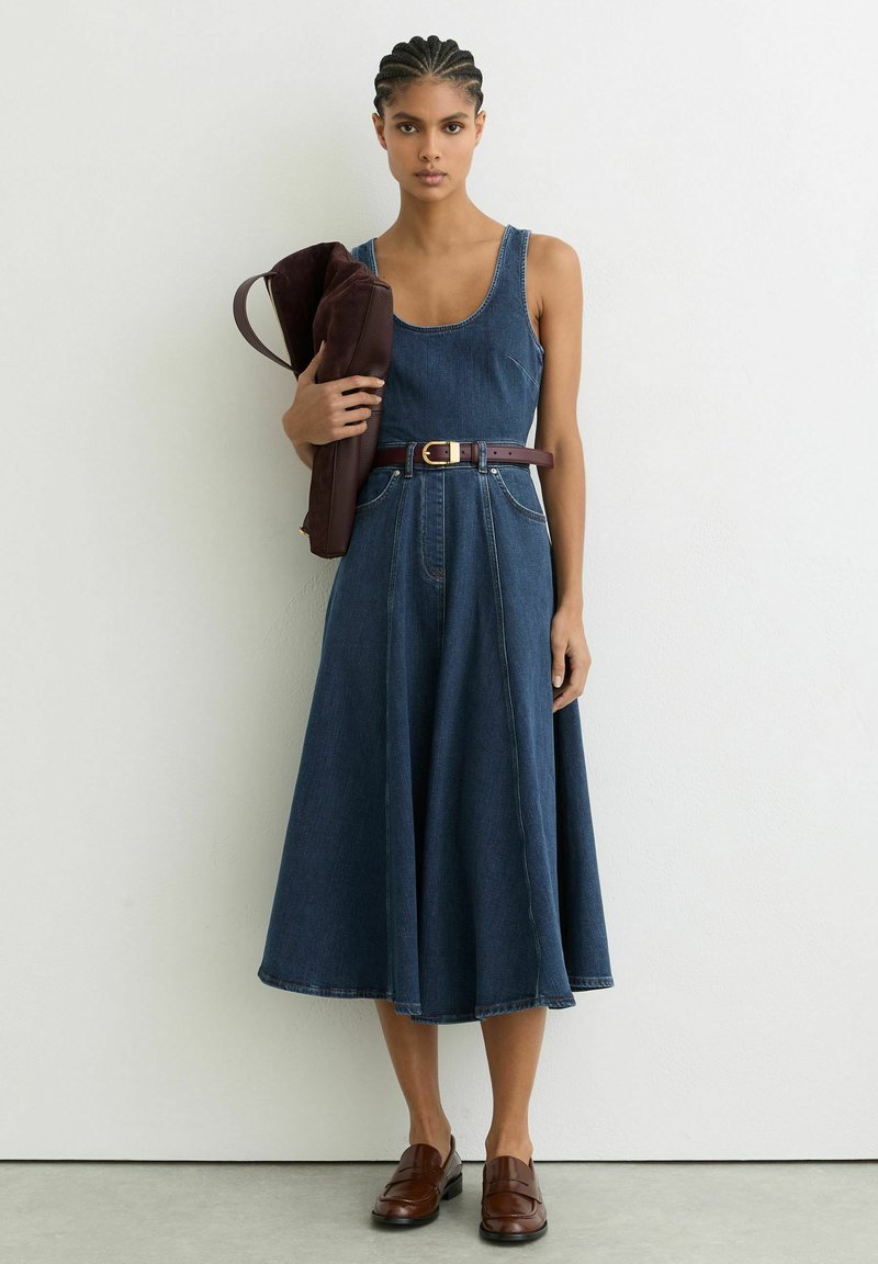 Kobieta z warkoczami w sukience midi z denimu bez rękawów i brązowym paskiem, trzyma złożoną brązową skórzaną torbę, stoi na tle białej ściany.