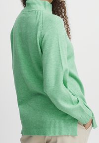 Pull vert en maille avec un col montant côtelé. Comprend un ourlet côtelé et des fentes latérales, une texture douce et une coupe ample.