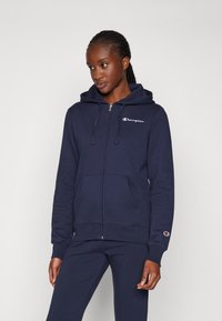 Navy blå zip-up hettegenser laget av myk fleece. Har frontlommer, snor i hetta, og brodert logo på brystet og ermet.