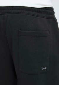 Schwarze Jogginghosen mit einer einfachen Gesäßtasche und einem Stoffetikett. Hergestellt aus weichem Material mit glatter Textur und elastischem Bund.