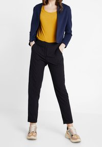 Meriini raidallinen bleiseri sinapinkeltaisen paidan yli, yhdistettynä mustiin raidallisiin housuihin ja monivärisiin tennareihin.