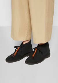 Schwarze Wildleder-Ankle-Boots mit runder Spitze und braunen Schnürsenkeln. Verfügt über eine flexible Gummisohle und ein minimalistisches Design.
