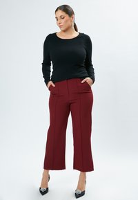 Schwarzes Langarmoberteil mit rundem Ausschnitt; burgunderrote, weit geschnittene Hose mit vorderen Nähten; das Model trägt schwarze High Heels mit dekorativen Akzenten.