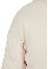 Lys beige puffjakke med en glatt, skinnende tekstur. Har brede kvilte seksjoner langs skuldrene for isolasjon og varme.