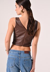Top corto in pelle marrone con uno scollo a V profondo e una zip visibile. Abbinato a jeans di denim azzurro chiaro con una toppa in pelle marrone.