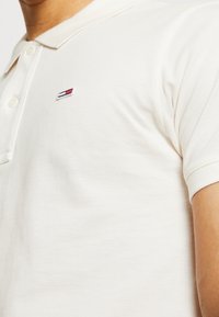 Polo de crema con mangas cortas, tejido texturizado, botonera de dos botones y un pequeño logo bordado con acentos en rojo, blanco y azul.