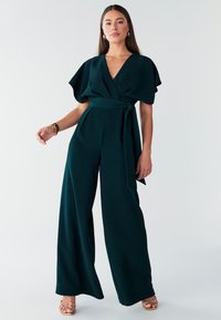 Tirkīzzils jumpsuits ar platiem kāju galiem, īsām piedurknēm un jostu viduklī. Gluda auduma ar vieglu spīdumu. Izskatu papildina zelta sandales.