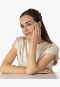 Goldring an einer Hand, die ein Gesicht hält. Das Subjekt trägt eine leichte Satinbluse. Glatte Haut, langes Haar, neutraler Hintergrund.