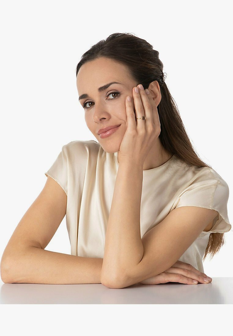 Goldring an einer Hand, die ein Gesicht hält. Das Subjekt trägt eine leichte Satinbluse. Glatte Haut, langes Haar, neutraler Hintergrund.