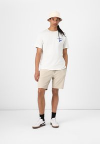 Conjunto informal que incluye una camiseta crema con un logo azul, pantalones cortos beige y un sombrero tipo bucket de tono claro; zapatillas blancas con suelas marrones y calcetines negros.