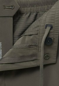 Gros plan sur un pantalon vert olive montrant un bouton, un cordon, un bouton-pression et un tissu intérieur avec un motif à carreaux et des détails cousus.