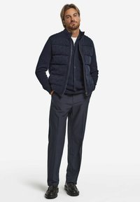 Granatowa pikowana kamizelka na granatowym kardiganie zapinanym na guziki, zestawiona z pasującymi spodniami i czarnymi eleganckimi butami. Tkanina wydaje się mieć teksturę i warstwową strukturę.