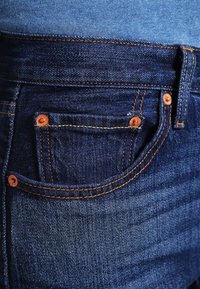 Tummansiniset farkut etutaskulla, joissa on kuparinvanteet ja oranssit tikkaukset saumoissa, luoden tekstuuri-ilmeen.