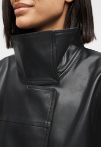 Γυναίκα που φοράει μαύρο δερμάτινο μπουφάν με ψηλό γιακά, ορατή ραφή και δομημένο, διπλωμένο σχέδιο.