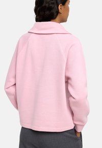 Różowy sweter z kołnierzykiem, o luźnym kroju, z rękawami raglanowymi i ściągaczami na rękawach. Gładka tekstura, jednolity kolor, minimalne detale.