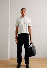 Weißes Poloshirt mit schwarzem Kragen, schwarze Hose und schwarze Schuhe. Model hält eine schwarze Lederhandtasche und steht vor einem hellen Hintergrund.