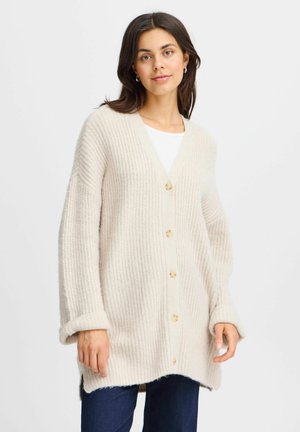 Cardigan beige lavorato a maglia con scollo a V, sette bottoni e maniche lunghe arrotolate. Presenta una texture a costine e un orlo posteriore più lungo.