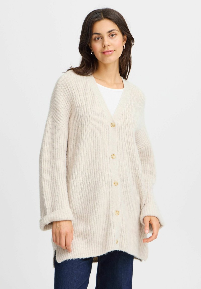 Cardigan beige lavorato a maglia con scollo a V, sette bottoni e maniche lunghe arrotolate. Presenta una texture a costine e un orlo posteriore più lungo.
