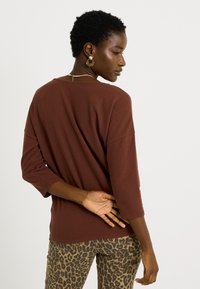 Camiseta marrón de manga larga con un corte holgado y cuello redondo, combinada con pantalones de estampado de leopardo. Presenta un detalle en la parte trasera del cuello.