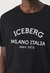 Musta puuvillapaita, jossa on valkoista tekstiä "ICEBERG MILANO ITALIA since 1974" lihavoituna, kaarevalla typografialla rintapielellä.