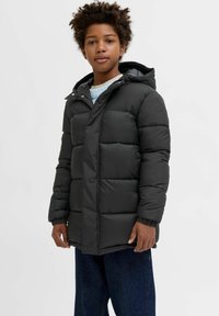 Melna pūkainā jaka ar kapuci, ar iegūtu šuvju dizainu un elastīgām piedurknēm. Uzvilkta virs gaiši zilas adītas kreklai un tumšiem džinsiem.