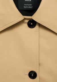 Manteau beige déperlant avec un col pointu et deux boutons noirs fermés sur le devant, étiquette indiquant la taille et l'origine.