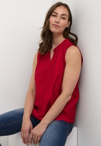 Piros, ujjatlan blúz V-nyakkal és gyűrt nyakrésszel, kék farmernadrággal viselve, semleges háttér előtt. Sima szövettextúra.
