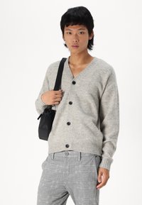 Helles graues Strickcardigan mit V-Ausschnitt, geripptem Saum und schwarzen Knöpfen. Kombiniert mit grauen karierter Hose und einer kleinen schwarzen Umhängetasche.