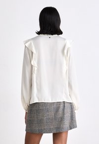 Ilgi piedurkņu ivora blūze ar volāniem uz pleciem, gluda tekstūra un savākta apkakle, apvienota ar pelēku svītrainu svārku.