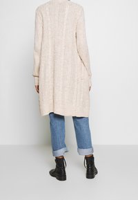 Lång, ljusbeige stickad cardigan med texturerade mönster, ribbade ärmslut och fåll. Bärs över uppvikt blå jeans och svarta ankelstövlar.