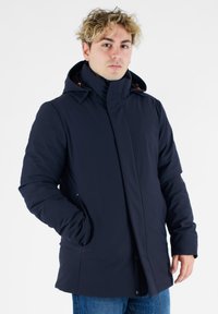 Marineblå isoleret parka med høj krave og hætte, der har lynlås og knaplukning, sidelommer og kontrastfarvede orange detaljer.
