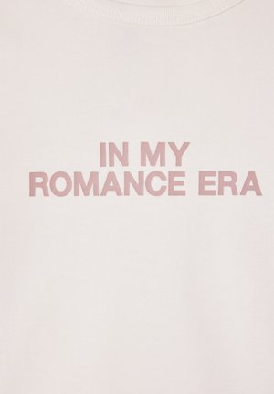 T-shirt in cotone rosa chiaro con scritte in rilievo di un rosa più scuro che recitano "NELLA MIA ERA ROMANTICA." Design semplice con scollo rotondo.