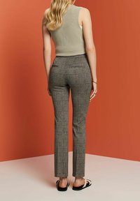 Femme aux longs cheveux blonds portant un haut en maille beige sans manches, un pantalon slim à carreaux taille haute et des sandales noires, debout contre un mur orange.