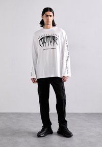 Balts garās piedurknes krekls ar izteiksmīgu melnu grafisko tekstu un ķēdes dizainu uz piedurknēm, apvienots ar melniem svītrainiem kravas biksēm un melnajām kedām.