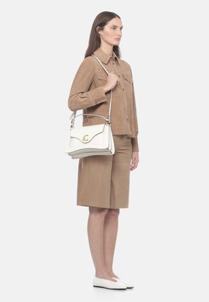 Woman wearing tan button-up jacket and matching knee-length shorts, holding white leather handbag with gold clasp, wearing white flats.