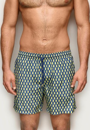 Uomo con pantaloncini da bagno blu con motivo di orsacchiotti gialli, in piedi con le braccia rilassate ai lati.
